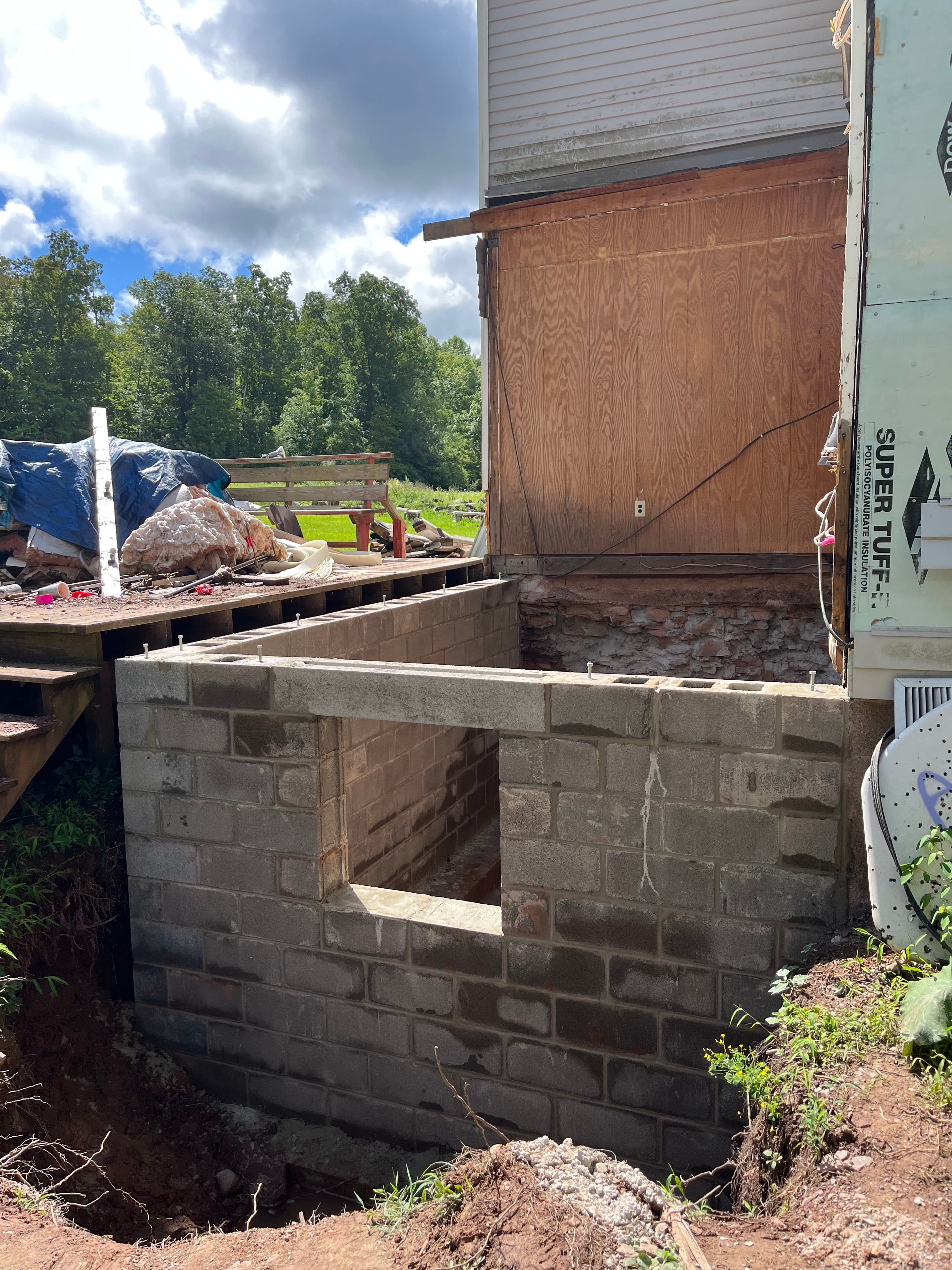 Concrete block foundation with a square opening under construction next to a plywood-sided building.