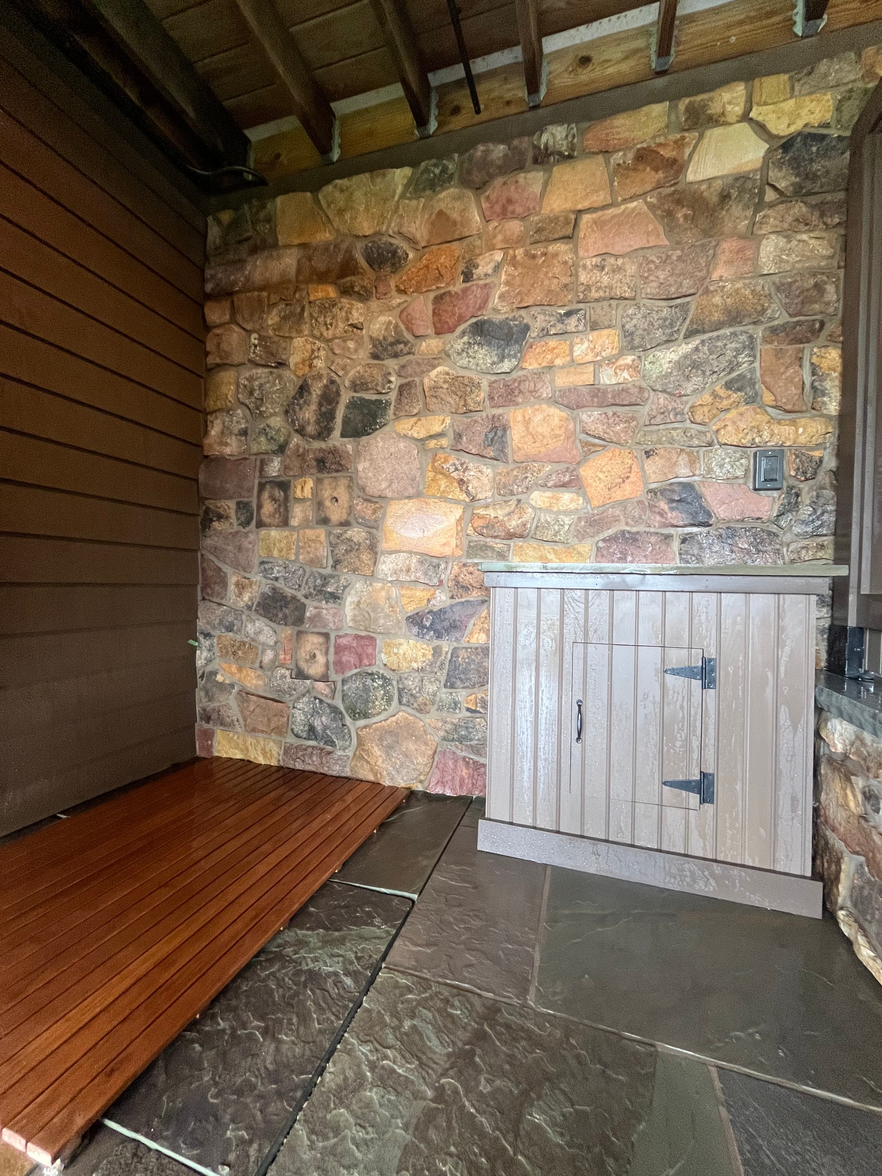 Small light brown access door set into a multi-colored stone wall above a brown deck.