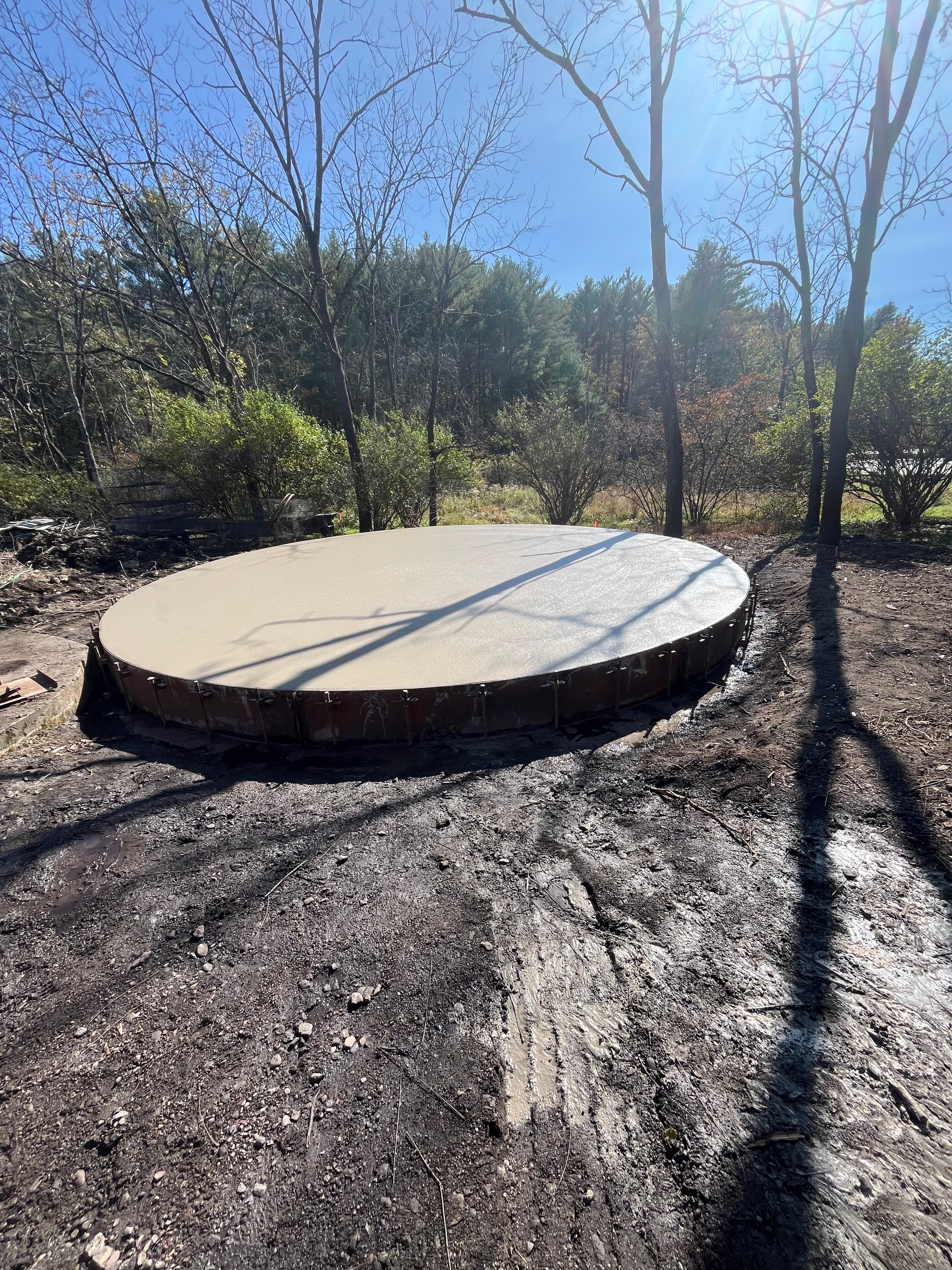 Circular concrete foundation pad in a muddy forest clearing with shadows from bare trees.