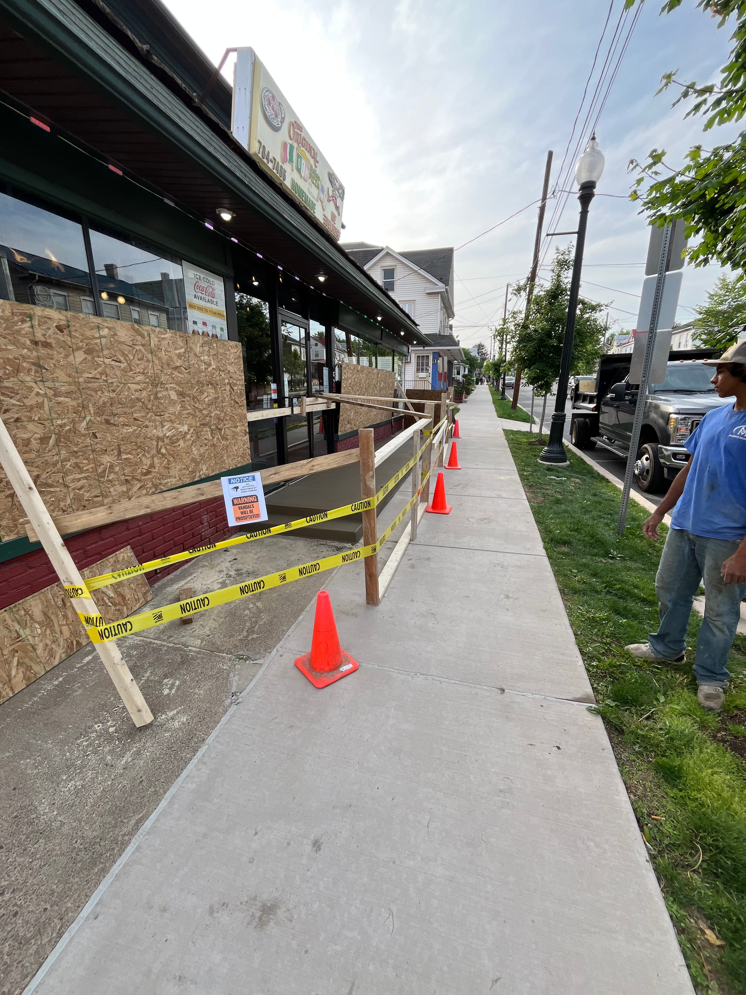 Boarded-up storefront windows behind yellow caution tape and orange cones on a concrete sidewalk.