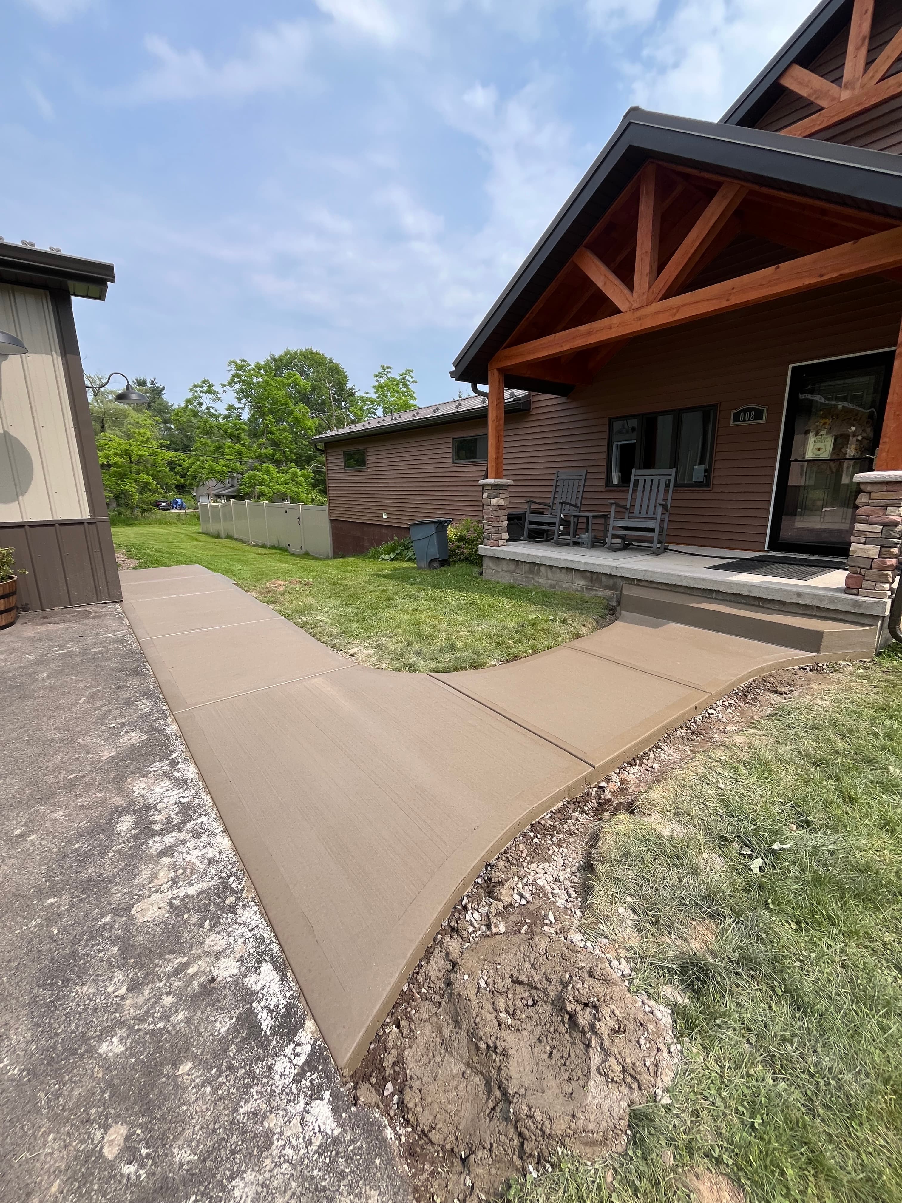Newly poured concrete walkway curving towards the wooden porch of a brown-sided house.