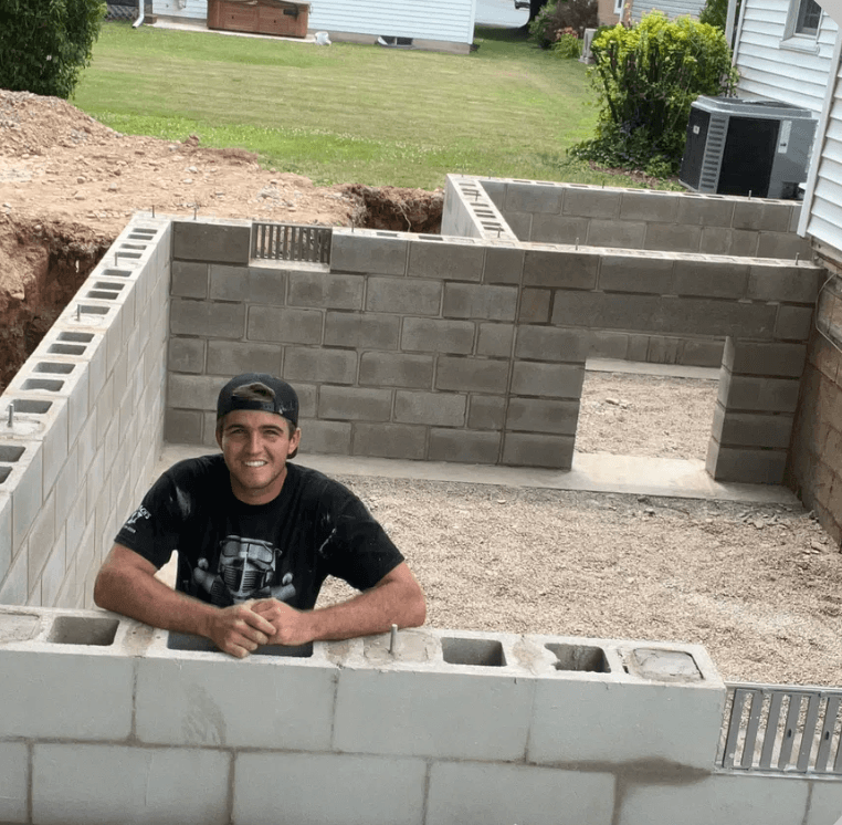 Smiling man leans on a concrete block wall foundation being built in a backyard.