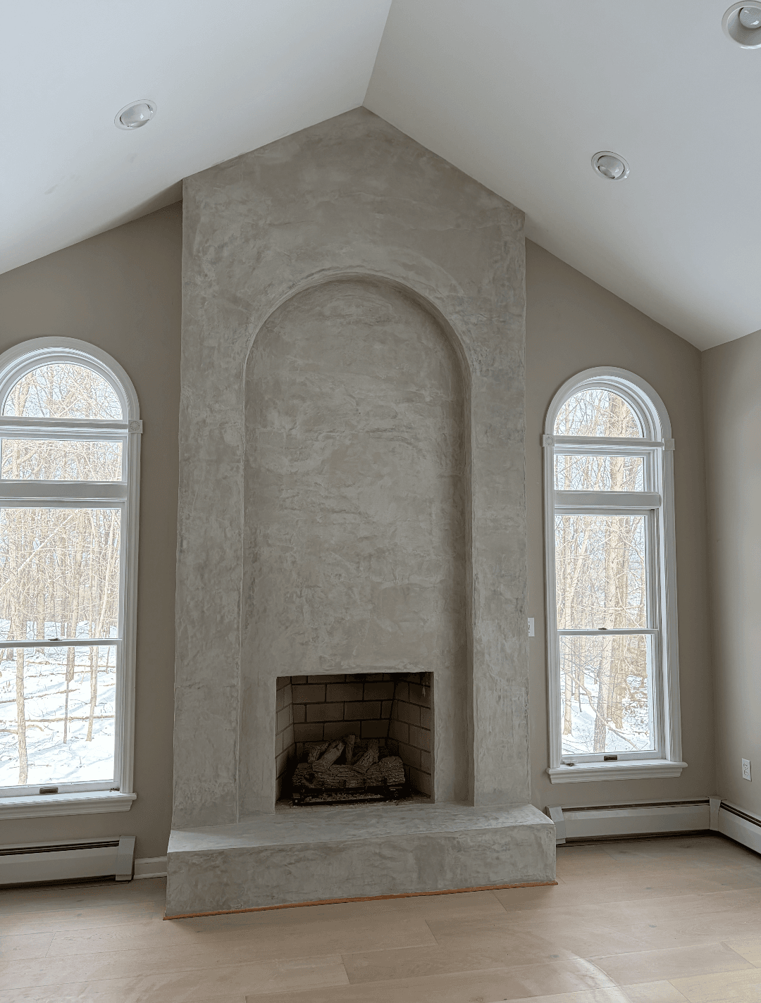 Tall gray concrete fireplace with an arched niche, flanked by windows overlooking snowy woods.